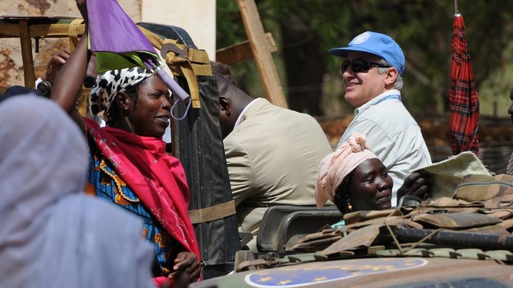 People gather near a military vehicle with UN markings; colorful fabrics and equipment are visible in the scene.