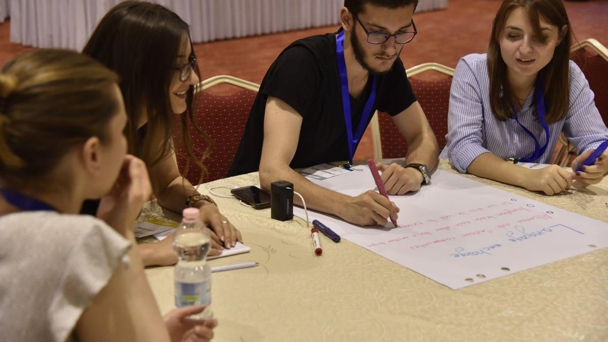 Four people seated around a table in a conference or workshop setting, collaborating on a large sheet of paper with handwritten notes. One person is writing with a pink marker while others observe and hold pens. Items on the table include a water bottle, markers, and a small black device. Red cushioned chairs and a long table with a white tablecloth are visible in the background.