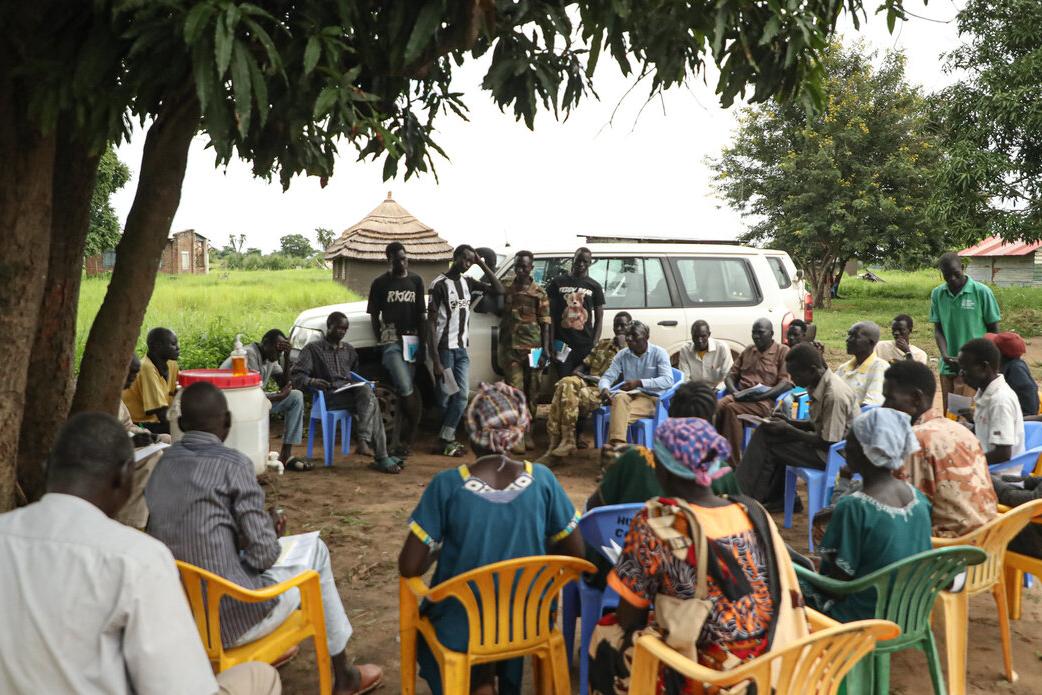 Community meeting held outdoors under a tree, with people seated in a circle on plastic chairs and several individuals standing near a white vehicle in the background.