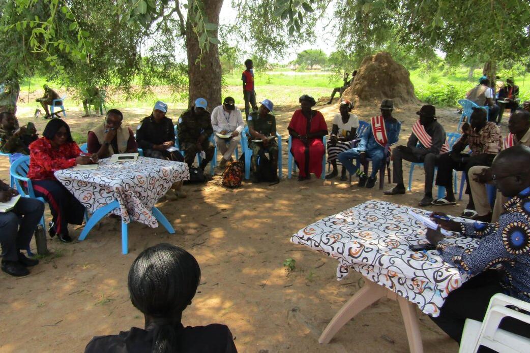 Group of people seated in a circle outdoors under a large tree, with two tables covered in patterned cloth at the center. Some participants wear blue caps, and traditional huts are visible in the background.