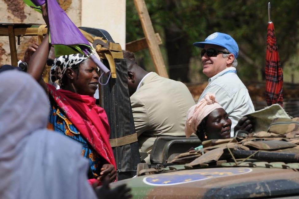 People gather near a military vehicle with UN markings; colorful fabrics and equipment are visible in the scene.