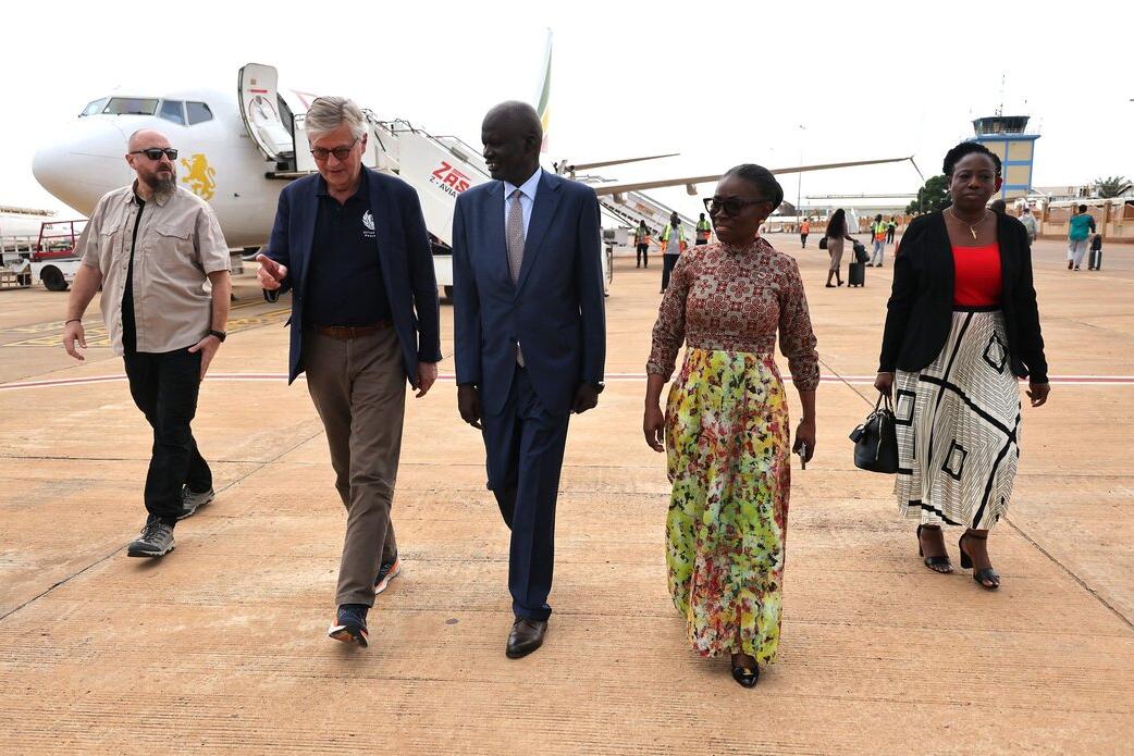 Four people including Under-Secretary-General Jean-Pierre Lacroix walking on an airport tarmac near a parked airplane, with other travelers and airport staff visible in the background.