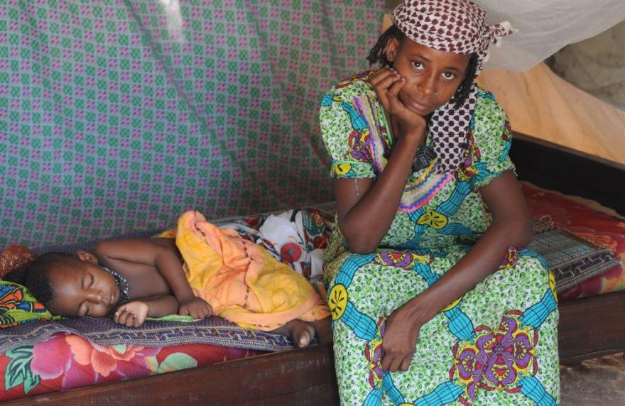 A UN-marked vehicle is parked on a dirt road with trees and buildings in the background