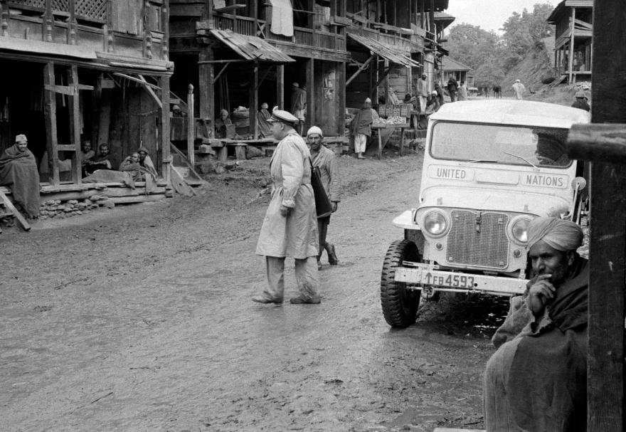 UN jeep parked on a muddy street in a village with wooden buildings. A UN officer in a coat walks nearby as locals sit and stand along the roadside.